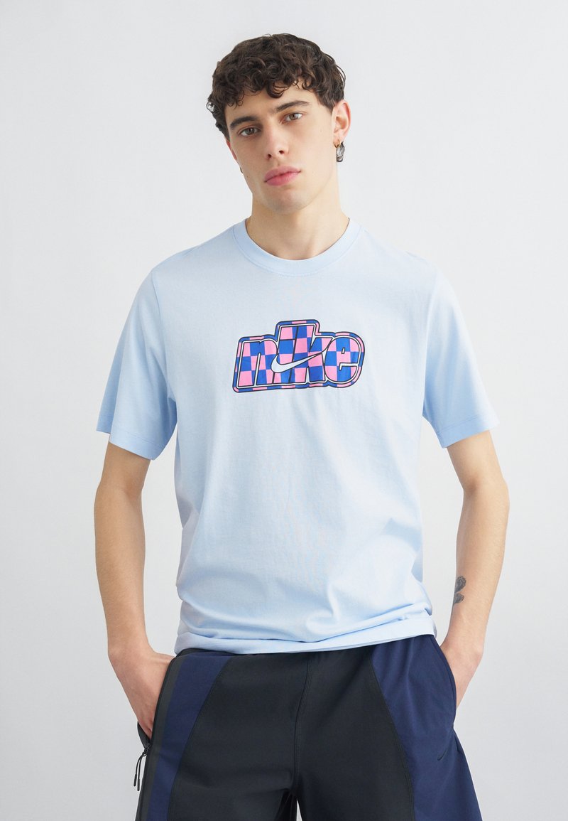 Mladenič s kodrastimi temnimi lasmi, ki nosi svetlomodro majico Nike z roza in modrim karo logotipom ter črne kratke hlače, stoji z rokami v žepih.