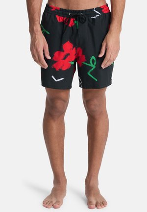Homme portant un short de bain noir avec des motifs floraux rouges et des feuilles vertes, debout pieds nus sur fond blanc.