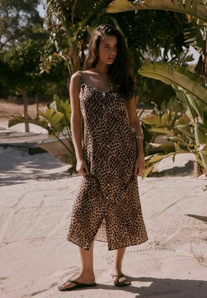 Žena s dlouhými tmavými vlasy, oblečená v bezrukávových šatech na knoflíky s leopardím vzorem a žabkách, stojící na písčité stezce poblíž velkých zelených rostlin.