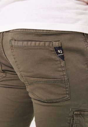 Los pantalones verde oliva presentan un bolsillo trasero con costura de marca, costuras visibles y una tela ligeramente texturizada.