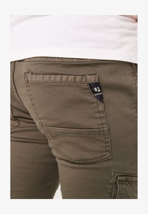 I pantaloni verde oliva presentano una tasca posteriore con cucitura brandizzata, cuciture visibili e un tessuto leggermente strutturato.