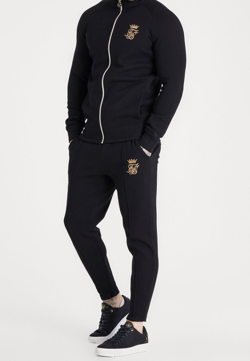 Giacca nera con zip e pantaloni jogger abbinati in tessuto morbido. Presentano dettagli del logo dorato e coulisse. Completato con scarpe da ginnastica nere.