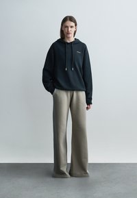 O femeie stă cu fața înainte, purtând un hanorac negru și pantaloni bej largi, pe un fundal gri simplu.