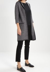 Abrigo gris de espiga con capucha, de apertura frontal y dos bolsillos laterales, combinado con una blusa negra y pantalones negros de corte ajustado, junto con unos zapatos planos.