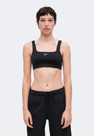 Mladá osoba s krátkymi tmavými vlasmi, oblečená v čiernu športovú podprsenku Nike a čierne nohavice so šnúrkou, stojaca na jednoduchom pozadí.