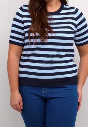 Femme portant un t-shirt à manches courtes à rayures horizontales bleu marine et bleu clair, associé à un jean bleu, debout devant un fond uni.