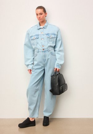 Młoda kobieta stojąca przy ścianie, ubrana w jasnoniebieską, oversize'ową kurtkę dżinsową i spodnie, trzymająca czarny skórzany plecak w prawej ręce.