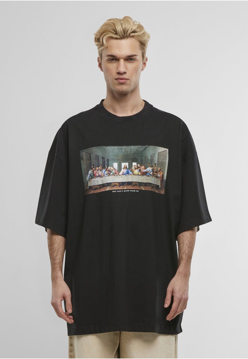 Ξανθός άντρας φοράει υπερμεγέθη μαύρο T-shirt με την εικόνα του Μυστικού Δείπνου και το κείμενο "ΔΕΝ ΜΠΟΡΕΙΣ ΝΑ ΚΡΕΜΑΣΕΙΣ ΜΑΖΙ ΜΑΣ" μπροστά.