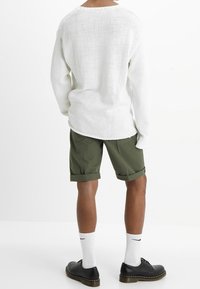 Bílý pletený svetr s volným střihem, doplněný olivově zelenými šortkami. Model má na sobě černé boty a bílé ponožky s logem.