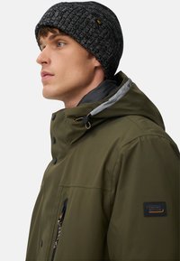 Grüne, wasserdichte Jacke mit Kapuze, ausgestattet mit schwarzen Knöpfen und einem grauen Innenfutter. Das Modell trägt eine schwarze, gerippte Mütze mit einem Logo.