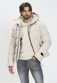 Svijetlosmeđi puffer jakna s kapuljačom, s zatvaranjem na zip i crnim detaljima, nosi se preko svijetlosmeđeg dukserice i sivih traperica. Meka tekstura.
