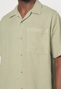 Chemise à manches courtes vert clair en tissu doux, avec un devant boutonné, un col cranté et une poche poitrine unique.