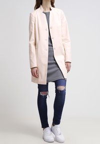 Abrigo largo de color rosa claro con un cuello simple y cinco botones, llevado sobre un vestido gris, combinado con jeans oscuros rasgados y zapatillas blancas.