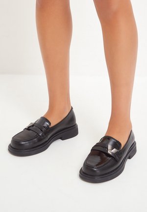 Jambes portant des mocassins en cuir noir brillant avec une semelle épaisse et un détail de boucle métallique, debout sur un sol blanc.