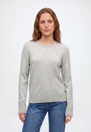 Vrouw met golvend lichtbruin haar draagt een lichtgrijze trui met ronde hals en blauwe spijkerbroek, staand tegen een effen witte achtergrond.