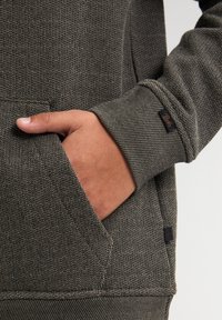 Felpa grigia con texture, dotata di tasca frontale, polsini a coste e un piccolo etichetta del marchio, che mette in evidenza un motivo tessuto per un tocco di dettaglio in più.