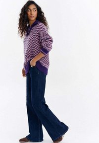 Kobieta o kręconych włosach, ubrana w fioletowy sweter w wzory, ciemnoniebieskie dzwoniaste jeansy oraz brązowe buty, stojąca z jedną ręką w kieszeni.