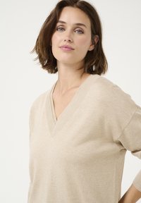 Pulover bej cu decolteu în V, dintr-un material moale, cu croială lejeră și umeri lăsați, având o textură subtilă și un design curat, minimalist.