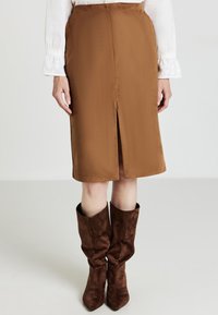Gonna marrone al ginocchio con uno spacco frontale, realizzata in tessuto liscio. Abbinata a stivali alti di suede marrone con effetto scivolato.