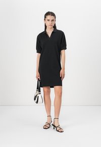 Black polo dress with short sleeves and a structured collar. Smooth fabric, knee-length design. Paired with strappy black sandals and a handbag.
