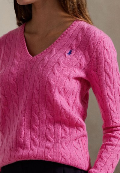 V-hals kabelstrikket genser i knallrosa ull, med en liten blå logo på brystet. Figurnær passform med ribbestrikkede mansjetter og kant.