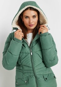 Mörkgrön puffjacka med en huva klädd i fuskpäls, dragkedja framtill och fickor med tryckknappar; har kviltade detaljer och ribbade manschetter.