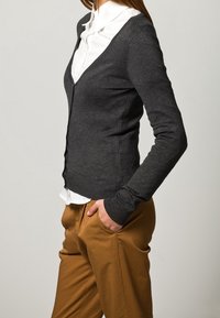 Grauer Wollcardigan mit V-Ausschnitt, gerippten Bündchen und taillierter Silhouette, über einem weißen Hemd mit Kragen getragen und mit beigen Hosen kombiniert.