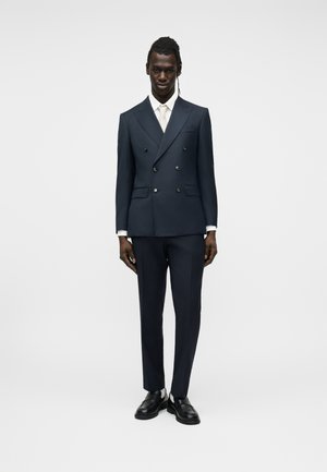 Bărbat în picioare, purtând un costum dublu pieptănat închis la culoare, cămașă albă, cravată albă și pantofi negri, pe un fundal simplu.