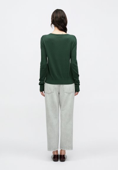 Πράσινο απαλό πλεκτό πουλόβερ με φαρδιά λαιμόκοψη και μακριά μανίκια, συνδυασμένο με ανοιχτό γκρι τζιν κανονικής γραμμής και απλά σκούρα παπούτσια.