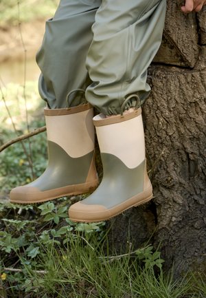 Kind trägt grüne und beige wasserdichte Stiefel und Hose, sitzt auf einem Baumstamm in einer grasbewachsenen Freiluftumgebung.