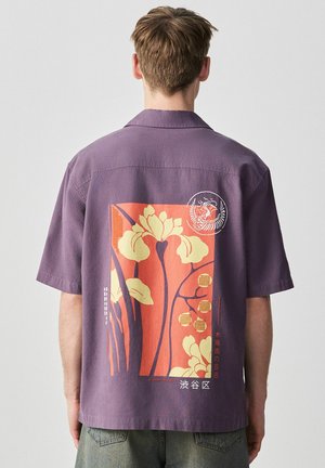 Man wearing purple short-sleeve shirt with orange and yellow floral graphic and Japanese text on back, standing against plain gray background.