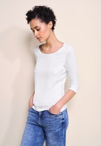 Hvid langærmet top med rund halsudskæring og afslappet pasform, parret med lyseblå jeans. Glat tekstur, minimal detaljering.