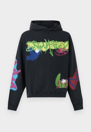 Jeune homme debout, portant un sweat à capuche noir et un jogging avec des imprimés colorés style graffiti, baskets vertes, sur fond clair uni.
