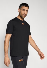 Μαύρο κοντομάνικο μπλουζάκι από λείο ύφασμα, με πορτοκαλί λογότυπο στο στήθος και λαιμόκοψη τύπου crew. Το κόψιμο είναι άνετο.