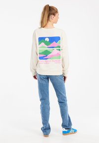 Sweat-shirt beige à manches longues avec un graphique coloré de paysage montagneux au dos, accompagné du texte "CE MOMENT MAGIQUE PROTESTATION DU LEVER DE SOLEIL TÔT."