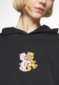 Black hooded sweatshirt featuring a front embroidered design of two cartoon bears, one pink and one yellow, embracing, with a heart detail.
