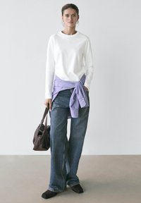 Woman in white long-sleeve shirt, lilac shirt tied at waist, wide-leg jeans, black shoes, holding dark brown handbag against plain background.