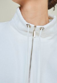 Giacca bianca con zip e colletto alto, cerniera argento e lacci bianchi, indossata da una persona con collo e mento visibili.