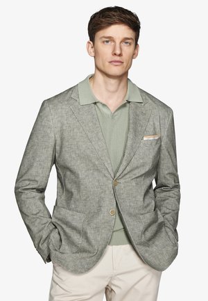 Uomo che indossa un blazer grigio chiaro con trama sopra una polo in maglia verde salvia e pantaloni beige con le mani in tasca su sfondo bianco.