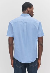 Chemise bleu clair à manches courtes en mélange de coton texturé avec un col boutonné et un pli au dos, associée à un jean en denim foncé.