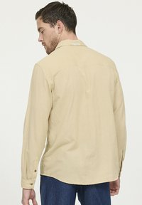 Beige langslavende shirt met een kraag, achterste yoke-detail, knoopmanchetten en een gebogen onderkant, gemaakt van zachte stof met een subtiele textuur.