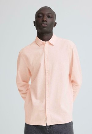 Ung mand iført en lyserød skjorte med knapper og lange ærmer, stående med hænderne bag ryggen mod en ensfarvet baggrund.
