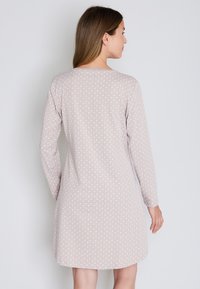 Donna con i capelli castani che indossa un vestito lungo fino al ginocchio, a maniche lunghe, di un beige chiaro con un piccolo motivo geometrico bianco, di spalle.