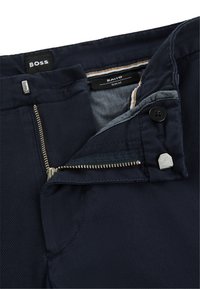 Navy slim-fit trousers feature a textured fabric with a zippered fly, button closure, and branded label. Metallic hardware adds detail.