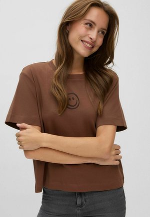 Femme aux longs cheveux bruns souriante, portant un t-shirt marron avec un petit visage souriant et un pantalon gris foncé, les bras croisés.