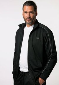 Chaqueta deportiva negra con cremalleras, con acentos grises en los laterales, fabricada en un tejido suave, con cuello alto y dos bolsillos delanteros, llevada sobre una camiseta blanca.