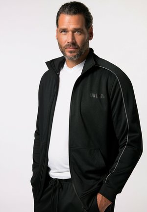 Chaqueta deportiva negra con cremalleras, con acentos grises en los laterales, fabricada en un tejido suave, con cuello alto y dos bolsillos delanteros, llevada sobre una camiseta blanca.