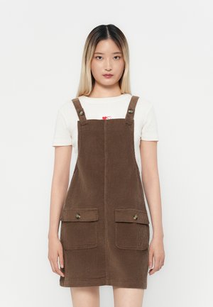 Bruine corduroy tuinbroek met schouderbanden, twee zijzakken en ronde knopen. Draagt over een wit shirt met een hartafbeelding.