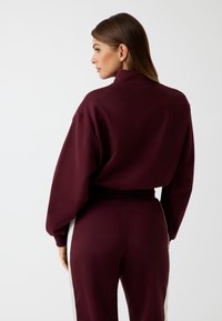 Set di abbigliamento attivo in bordeaux, composto da una felpa a collo alto con spalle basse e pantaloni coordinati con strisce bianche laterali. Texture morbida.