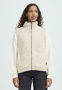 Gilet in pile beige con collo alto, chiusura con zip completa, due tasche laterali e una superficie morbida e testurizzata, indossato sopra un maglione crema.